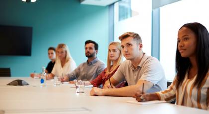 Group of people sat along table listening to speaker